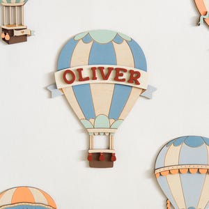 Pode incluir: Decoração de parede de balão de ar quente de madeira em azul, bege e verde-azulado. O nome "OLIVER" é exibido em uma faixa. Outros balões semelhantes são visíveis ao fundo, criando um tema de berçário lúdico.
