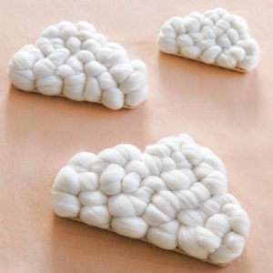 May include: Three white cloud-shaped decorative items. Each cloud is made of a textured, woven material, giving a soft, puffy appearance. The clouds are arranged on a light pink surface, creating a whimsical and cozy aesthetic.