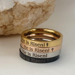 May include: Four stacked stainless steel rings with the inscription "He is Risen!" and a cross symbol on each ring. The rings are in gold, rose gold, silver, and black.