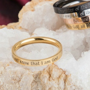 Op de afbeelding: Een set van vijf stapelbare ringen in goud, zilver en zwart met gegraveerde inspirerende boodschappen. De ringen bevatten de zinnen "Dochter van de Koning", "Dochter van de Soort", "Gezegend", "Met God is alles mogelijk" en "en weet dat ik God ben".