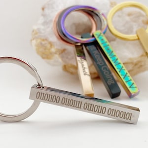 Op de afbeelding: Een set van vijf zilveren sleutelhangers met verschillende gegraveerde ontwerpen. De sleutelhangers liggen op een wit oppervlak. De ontwerpen omvatten "Keep Going", "God", "Blessed", Romeinse cijfers en een binaire code.