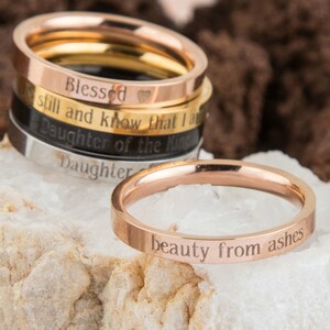 May include: A stack of five thin gold, silver, and black rings with engraved inspirational messages, including "Blessed", "still and know that I am", "Daughter of the King", and "Daughter". The bottom ring is rose gold and reads "beauty from ashes".