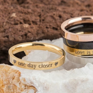 Könnte beinhalten: Ein Satz von fünf Ringen aus Gold, Silber und Roségold mit gravierten Botschaften. Der oberste Ring ist aus Roségold und trägt die Aufschrift "Love is patient". Der zweite Ring ist schwarz und trägt die Aufschrift "Love is patient". Der dritte Ring ist aus Gold und trägt die Aufschrift "Love is patient". Der vierte Ring ist aus Silber und trägt die Aufschrift "Love is patient". Der unterste Ring ist aus Gold und trägt die Aufschrift "one day closer" mit einem Herz auf jeder Seite.