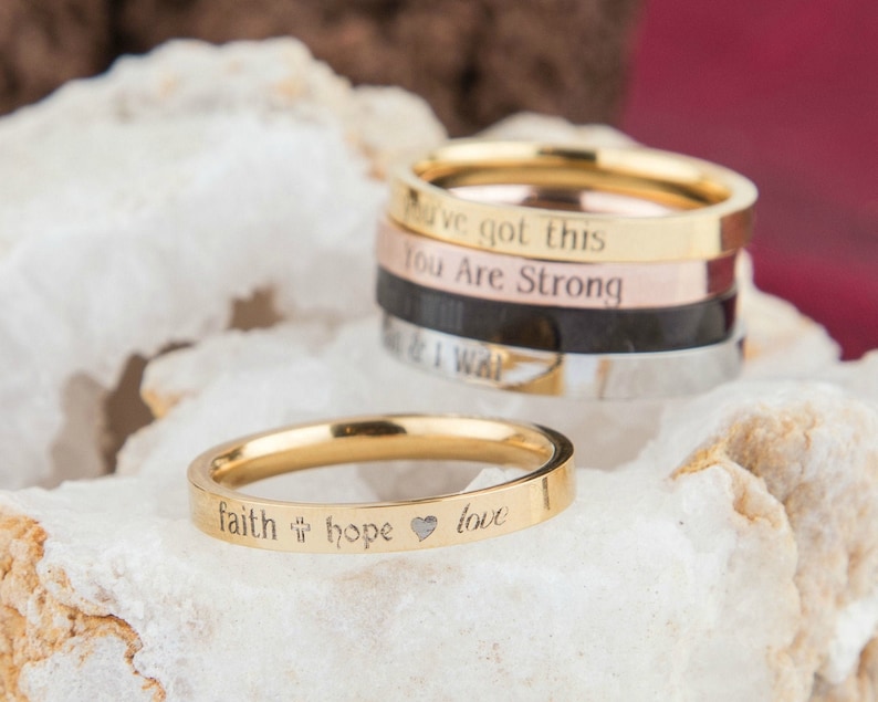May include: A set of five gold, silver, and rose gold stacking rings with engraved inspirational messages. The rings feature the phrases "You've got this", "You are strong", "Faith", "Hope", and "Love".