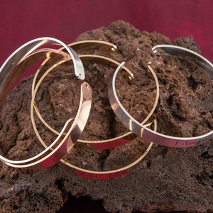 Puede incluir: Tres pulseras de brazalete apiladas, una plateada, una dorada y una de oro rosa. La pulsera plateada tiene la inscripci&oacute;n "I Am".