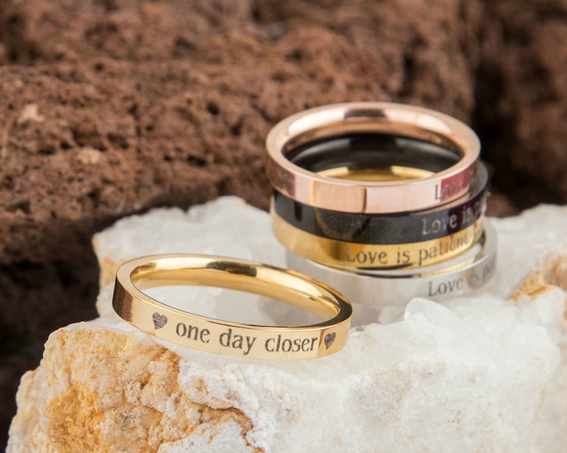Puede incluir: Un conjunto de cuatro anillos de oro, plata, negro y oro rosa con mensajes grabados. El anillo de oro tiene las palabras "one day closer" grabadas con un peque&ntilde;o coraz&oacute;n a cada lado del texto.