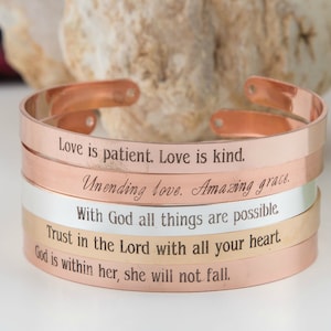 Könnte beinhalten: Fünf übereinander gestapelte Kupferarmbänder mit eingravierten inspirierenden Zitaten. Die Zitate lauten: "Liebe ist geduldig. Liebe ist gütig.", "Unendliche Liebe. Erstaunliche Gnade.", "Mit Gott ist alles möglich.", "Vertraue auf den Herrn von ganzem Herzen.", und "Gott ist in ihr, sie wird nicht fallen."