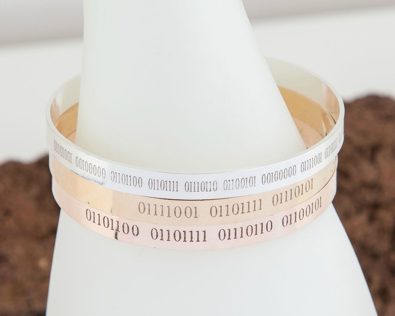 Puede incluir: Tres pulseras de plata con c&oacute;digo binario grabado. Las pulseras est&aacute;n apiladas una encima de la otra.