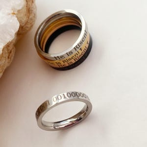 Op de afbeelding: Twee zilveren ringen. De bovenste ring is een stapel van drie ringen met de woorden "He is Risen" gegraveerd op de buitenband. De onderste ring heeft de nummers "000100100000" gegraveerd op de buitenband.