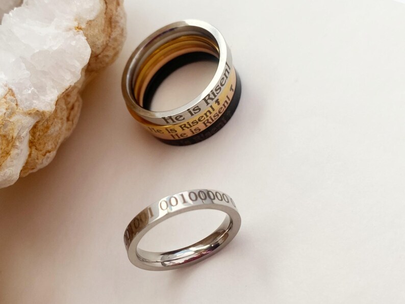 Op de afbeelding: Twee zilveren ringen. De bovenste ring is een stapel van drie ringen met de woorden "He is Risen" gegraveerd op de buitenband. De onderste ring heeft de nummers "000100100000" gegraveerd op de buitenband.