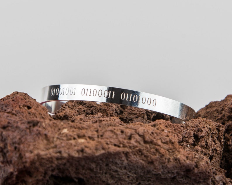 Puede incluir: Una pulsera de plata con el c&oacute;digo binario "01010001 01100011 01101000" grabado en ella. La pulsera est&aacute; apoyada en una roca marr&oacute;n.