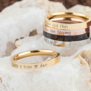 May include: Four gold, silver, rose gold, and black stacking rings with engraved inspirational messages. The top ring reads "You've got this". The second ring reads "You are strong". The third ring reads "Keep going". The bottom ring reads "I will". A single gold ring with the words "faith hope love" and a heart symbol is shown in the foreground.