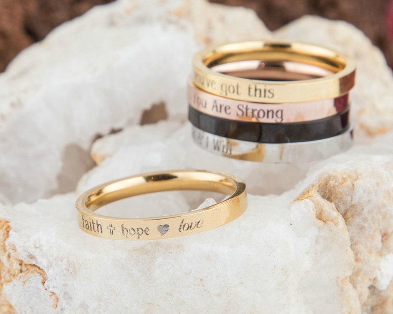 May include: Four gold, silver, rose gold, and black stacking rings with engraved inspirational messages. The top ring reads "You've got this". The second ring reads "You are strong". The third ring reads "Keep going". The bottom ring reads "I will". A single gold ring with the words "faith hope love" and a heart symbol is shown in the foreground.