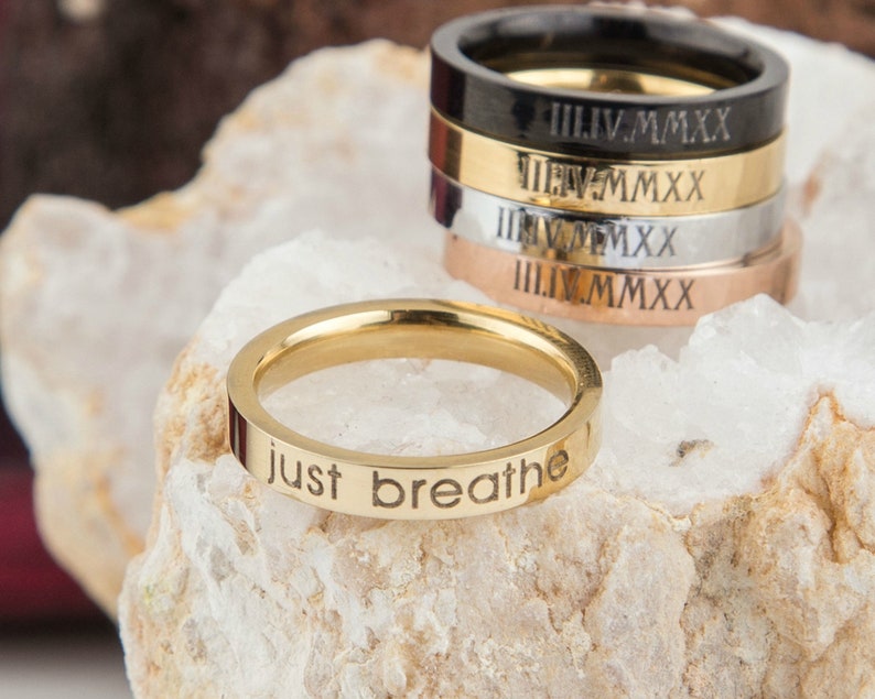 May include: A gold-toned ring with the words "just breathe" engraved on the band. The ring is sitting on a white rock. There are several other rings stacked behind it, some with Roman numeral engravings.