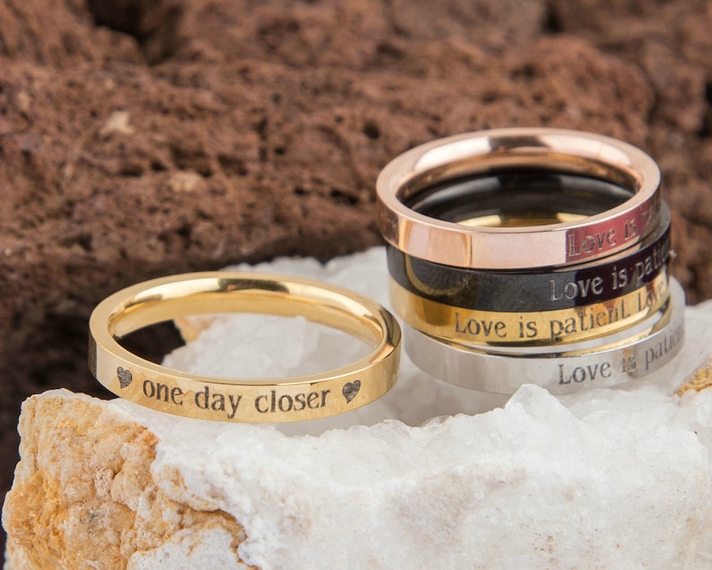 Puede incluir: Un conjunto de cinco anillos de oro, plata y oro rosa con mensajes grabados. El anillo superior es de oro rosa y dice "Love is patient". El segundo anillo es negro y dice "Love is patient". El tercer anillo es de oro y dice "Love is patient". El cuarto anillo es de plata y dice "Love is patient". El anillo inferior es de oro y dice "one day closer" con un coraz&oacute;n a cada lado.