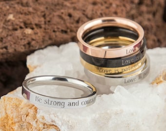 May include: A stack of rings in various colors, including silver, gold, black, and rose gold. One silver ring is separate, with the inscription "Be strong and courageous." Other rings have the words "Love is patient" and "Love is..." engraved.
