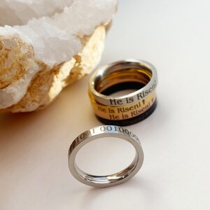 Op de afbeelding: Drie zilveren ringen die op elkaar gestapeld zijn. De bovenste ring heeft de woorden "He is Risen" gegraveerd. De middelste ring heeft de woorden "He is Risen!" gegraveerd. De onderste ring heeft de woorden "He is Risen!" gegraveerd. De ringen liggen op een wit oppervlak.