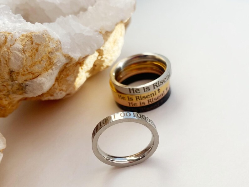Op de afbeelding: Drie zilveren ringen die op elkaar gestapeld zijn. De bovenste ring heeft de woorden "He is Risen" gegraveerd. De middelste ring heeft de woorden "He is Risen!" gegraveerd. De onderste ring heeft de woorden "He is Risen!" gegraveerd. De ringen liggen op een wit oppervlak.