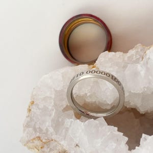 Op de afbeelding: Een zilveren ring met de inscriptie "10 0000 100" gegraveerd op de band. De ring ligt op een witte kristallen steen.