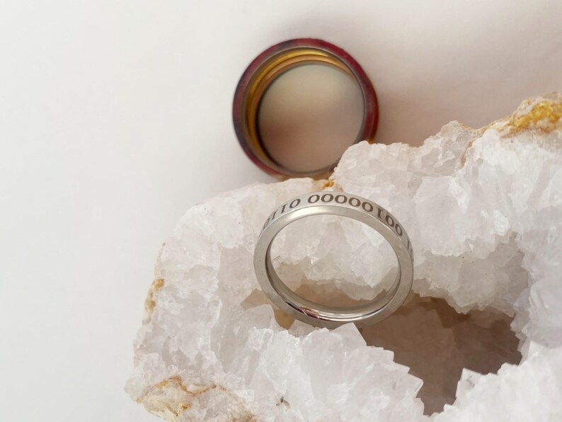 Op de afbeelding: Een zilveren ring met de inscriptie "10 0000 100" gegraveerd op de band. De ring ligt op een witte kristallen steen.