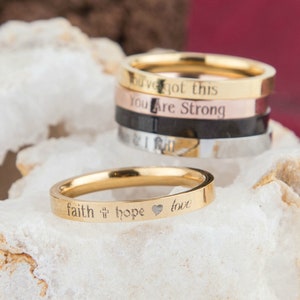 May include: A set of five gold, silver, and black stacking rings with engraved inspirational messages, including "You've got this", "You are strong", "Faith", "Hope", and "Love".