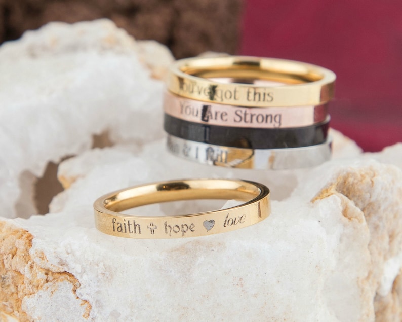 May include: A set of five gold, silver, and black stacking rings with engraved inspirational messages, including "You've got this", "You are strong", "Faith", "Hope", and "Love".