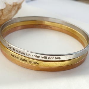 May include: Three stacked bangle bracelets in silver, gold, and rose gold. The silver bracelet has the inscription "God is within her; she will not fall." The gold bracelet has the inscription "custom date, quote".