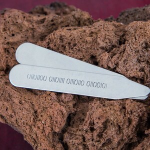 Può includere: Due ferri da colletto in argento con un motivo ripetuto della parola "OIOIOI" incisa sulla superficie. I ferri da colletto sono appoggiati su una superficie marrone testurizzata.