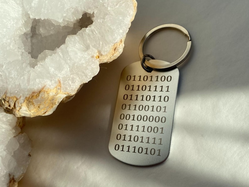 K&ouml;nnte beinhalten: Ein silberner Schl&uuml;sselanh&auml;nger mit einem rechteckigen Anh&auml;nger, der mit einer Reihe von Bin&auml;rcode graviert ist: 01101100, 01101111, 01110110, 01100101, 00100000, 01111001, 01101111, 01110101