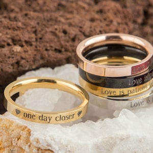 Puede incluir: Un conjunto de cuatro anillos de oro, plata, negro y oro rosa con mensajes grabados. El anillo de oro tiene la inscripci&oacute;n "one day closer" con un coraz&oacute;n a cada lado.