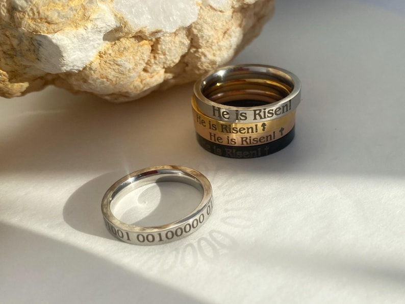 Op de afbeelding: Vier zilveren ringen gestapeld op elkaar. De bovenste ring heeft de woorden "He is Risen!" gegraveerd. De tweede ring heeft de woorden "He is Risen!" gegraveerd. De derde ring heeft de woorden "He is Risen!" gegraveerd. De onderste ring heeft de woorden "He is Risen!" gegraveerd. Een zilveren ring met de nummers "1001 00100000 01" gegraveerd.