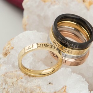 May include: A set of five gold, silver, and black rings with engraved roman numerals and the words "just breathe".