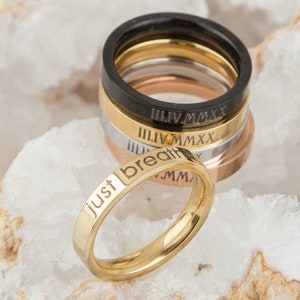 May include: A set of five gold, silver, and black stacking rings with Roman numeral engravings. The bottom ring is gold and engraved with the words "Just breath".