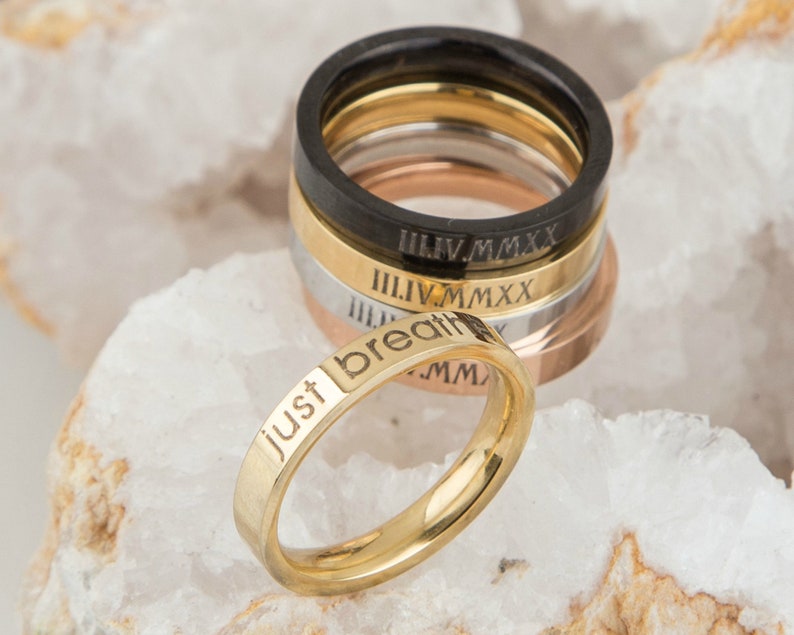 May include: A set of five gold, silver, and black stacking rings with Roman numeral engravings. The bottom ring is gold and engraved with the words "Just breath".