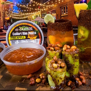 May include: A close-up of a jar of pineapple Colibri Rim Paste, a clear plastic container of orange sauce, and three green cucumber cups filled with peanuts and a red chili powder rim. The cups are on a wooden surface with more peanuts scattered around them.