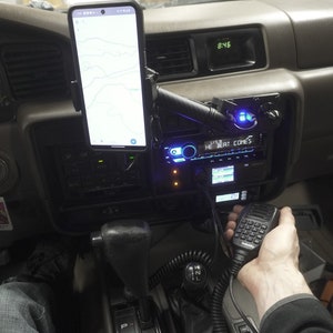 Puede incluir: Un salpicadero de coche negro con un teléfono inteligente en un soporte de teléfono, una radio con una pantalla azul y una radio de mano con un teclado negro y gris. La radio está conectada al salpicadero con un cable negro.