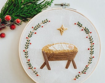 May include: Embroidered Christmas hoop art featuring a nativity scene. The design includes a baby in a manger, a star, and a garland of green leaves and red berries. The embroidery is set in a wooden hoop, with a sprig of greenery and berries in the background.