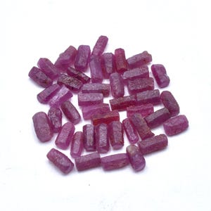 Peut inclure: Un ensemble de pierres précieuses de rubis brutes, affichant une gamme de teintes roses et rouges. Les pierres taillées grossièrement sont de forme irrégulière, avec des structures cristallines visibles. Les pierres sont dispersées sur une surface blanche.