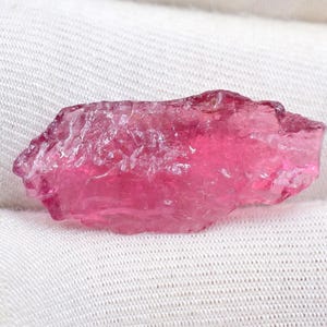 May include: A rough, translucent pink gemstone, possibly tourmaline, sits on white fabric. The stone has a crystalline structure and varying shades of pink, from light to deep magenta.