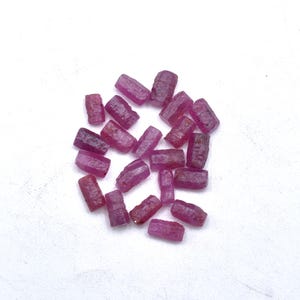 Pode incluir: Uma coleção de pedras preciosas de rubi bruto, cada uma com um tom vermelho-púrpura profundo. As pedras em bruto são retangulares e variam em tamanho, contra um fundo branco. As pedras preciosas não polidas mostram sua textura natural.