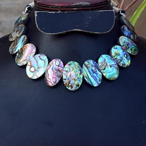 May include: A necklace featuring a series of oval-shaped abalone shell pieces. Each piece displays a unique iridescent pattern with swirling colors of blue, green, purple, and pink. The shell segments are linked together to form a curved collar.