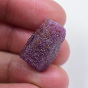 May include: A raw, uncut ruby gemstone with a deep magenta and purple hue. The stone features a rough, textured surface with natural crystalline facets and small golden-brown mineral inclusions scattered across its irregular, elongated shape.