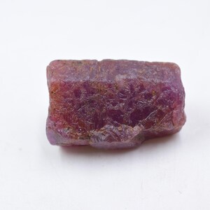 May include: A raw, unpolished ruby gemstone with a deep magenta and reddish-purple hue. The stone features a rough, textured surface with natural crystalline facets and irregular edges, displaying a translucent quality throughout its structure.