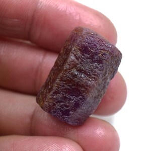 Pode incluir: Uma pedra preciosa de rubi bruta e não lapidada com um tom vermelho escuro e roxo. A pedra apresenta uma superfície rugosa e texturizada com facetas cristalinas naturais e uma aparência opaca, exibindo a sua forma irregular e não polida.