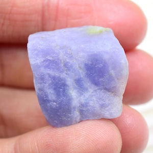 May include: A raw, unpolished piece of lavender-colored tanzanite gemstone with a rough, crystalline texture and subtle white veining throughout the stone. The mineral specimen displays a translucent quality with a pale yellow inclusion on the top edge.