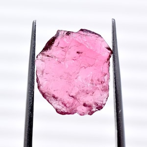 Könnte beinhalten: Ein roher, rosa Edelstein wird von einer Pinzette vor einem weißen Hintergrund gehalten. Der Edelstein hat ein durchscheinendes Aussehen mit einer unebenen Oberfläche. Die Farbe ist ein leuchtendes, mittleres Rosa mit Farbtonvariationen.