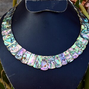 May include: A statement necklace composed of multiple rectangular abalone shell segments arranged in a curved, collar-style design. Each segment displays an iridescent, multi-colored surface featuring shimmering shades of green, blue, and pink.