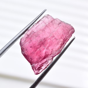 May include: A rough-cut, pink gemstone is held by a pair of silver tweezers against a white background. The gemstone has a crystalline structure and a vibrant, translucent color.