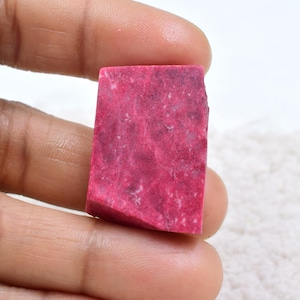 Pode incluir: Uma gema retangular, de corte bruto, em um tom magenta profundo. A pedra é segurada entre os dedos, mostrando sua superfície texturizada e variações de cor naturais. A gema parece ser um espécime bruto, não polido, ideal para fazer joias ou colecionar.
