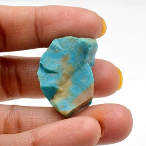 May include: A rough-cut turquoise gemstone with a mix of blue and tan hues. The stone has a natural, uneven texture and is held in a hand against a white background. The gemstone is a vibrant example of natural mineral beauty.